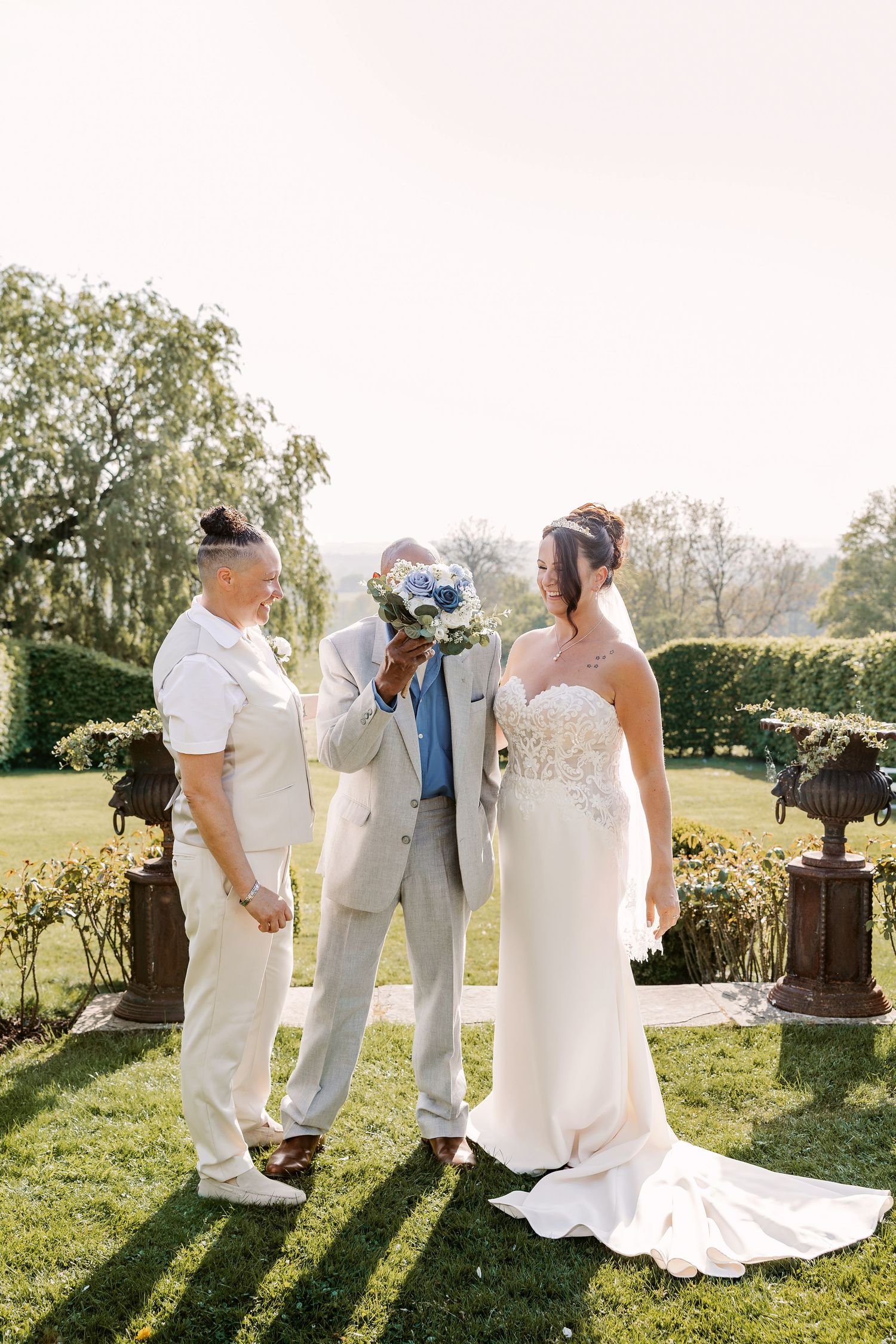 Wedding party dressed in white and cream attire poses together in a sunlit garden setting.