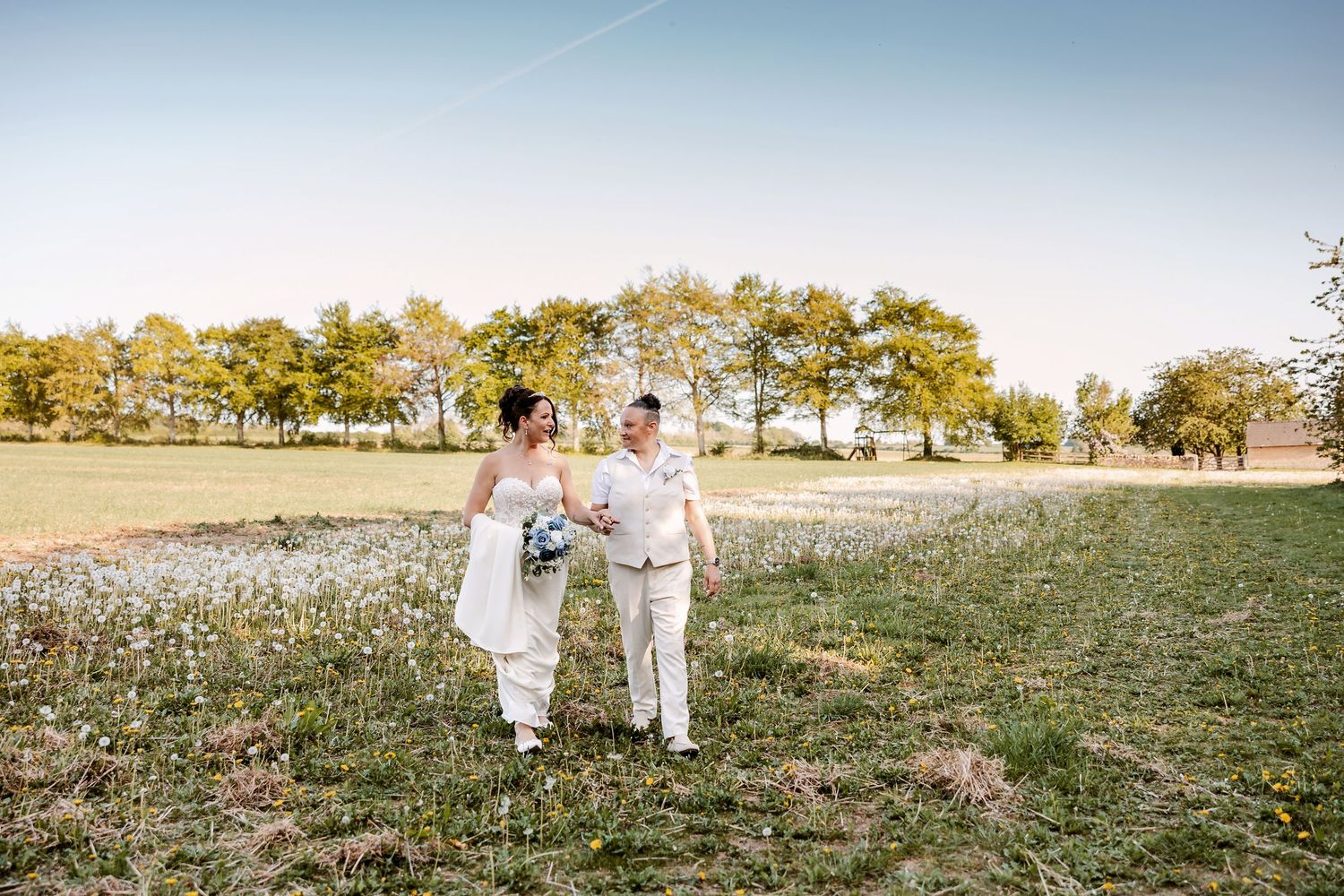 Wedding couple walks through sunny meadow with distant landscape views.