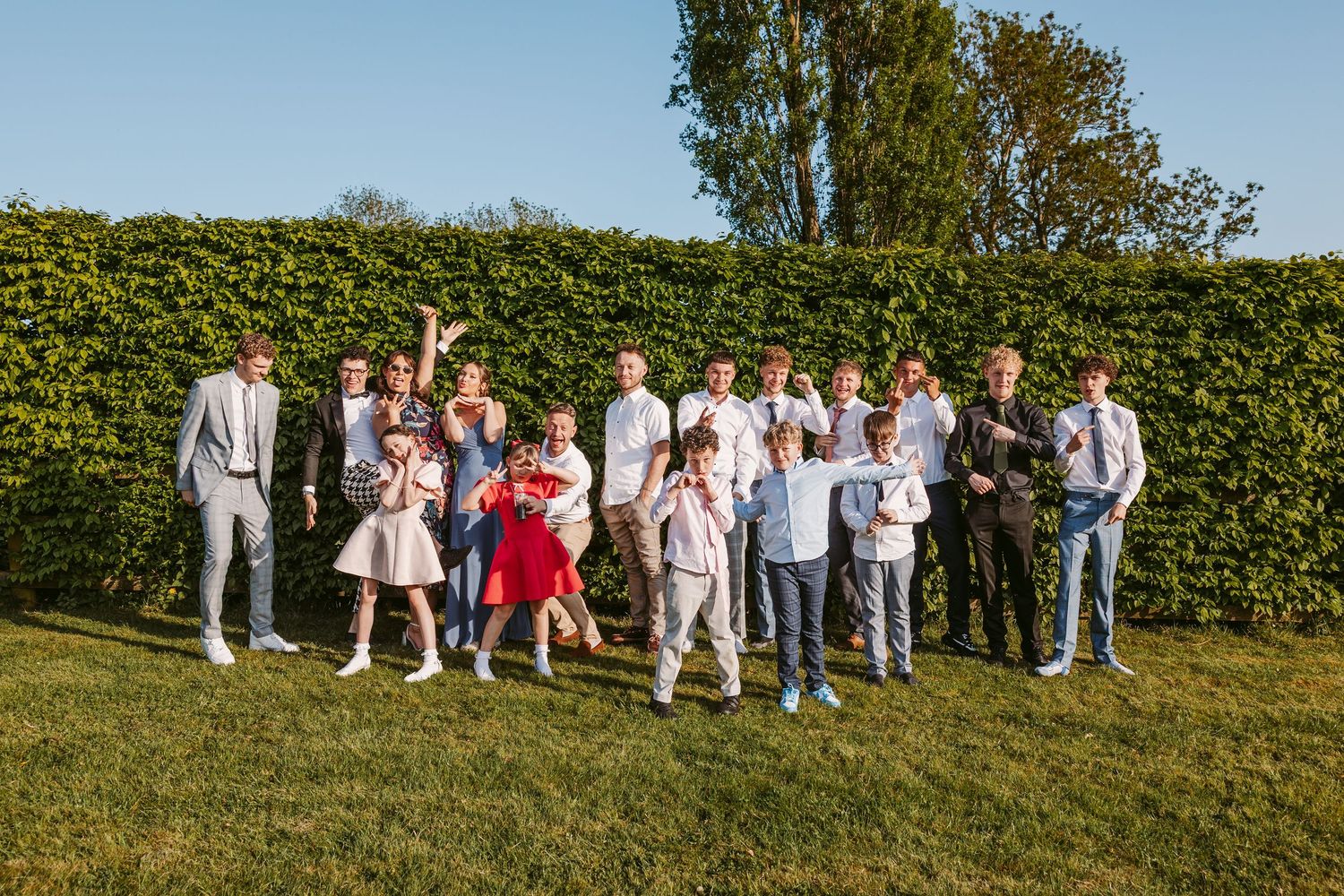 Wedding guests line up for a group photo against a lush green hedge backdrop on a sunny day.