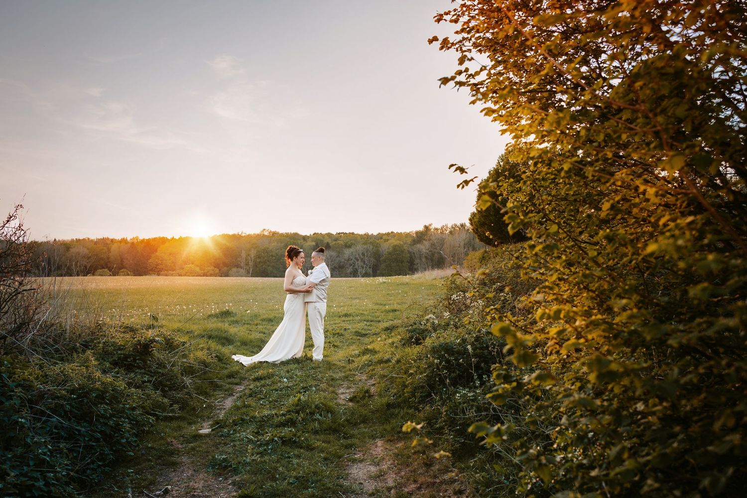 A couple in wedding attire stands in a golden meadow at sunset with autumn trees framing the romantic scene.