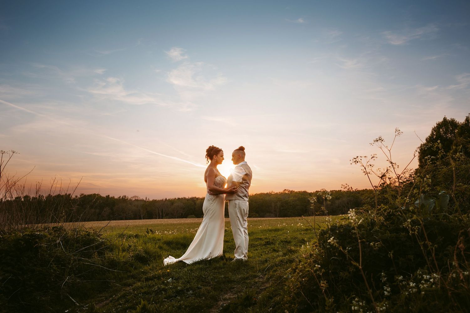 Sunset silhouettes a couple standing in a meadow against a golden sky with rolling hills in the background.