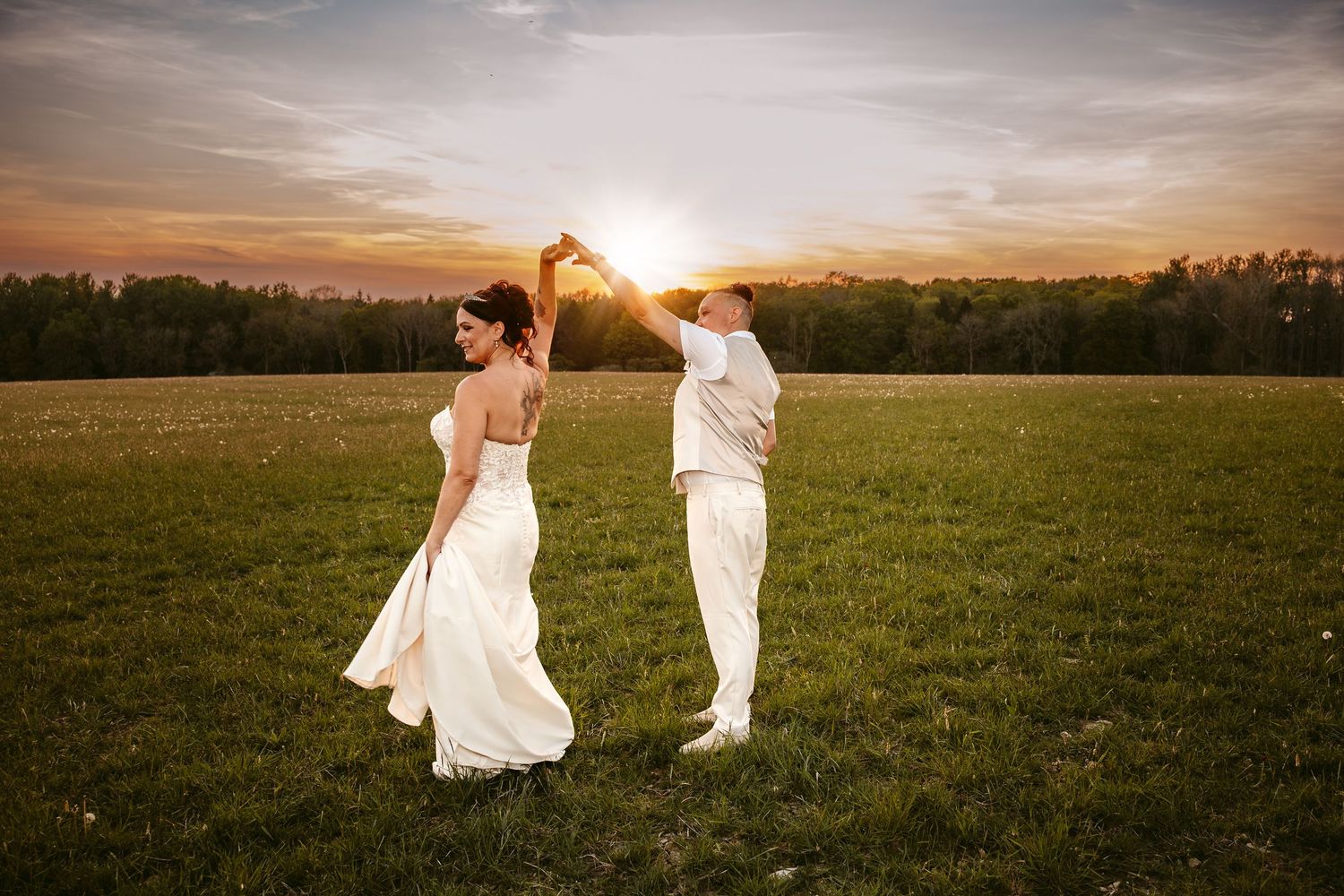 A couple dances together in a grassy field at sunset with golden sunlight streaming between them.