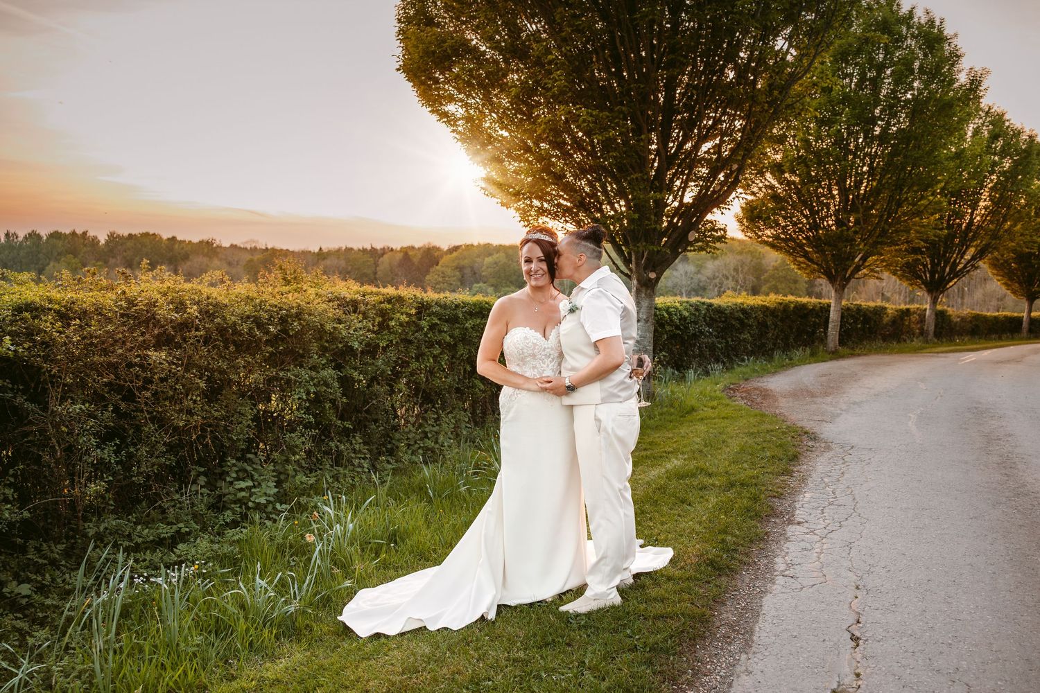 A couple in wedding attire poses together at sunset in a rustic outdoor setting with trees and a path.
