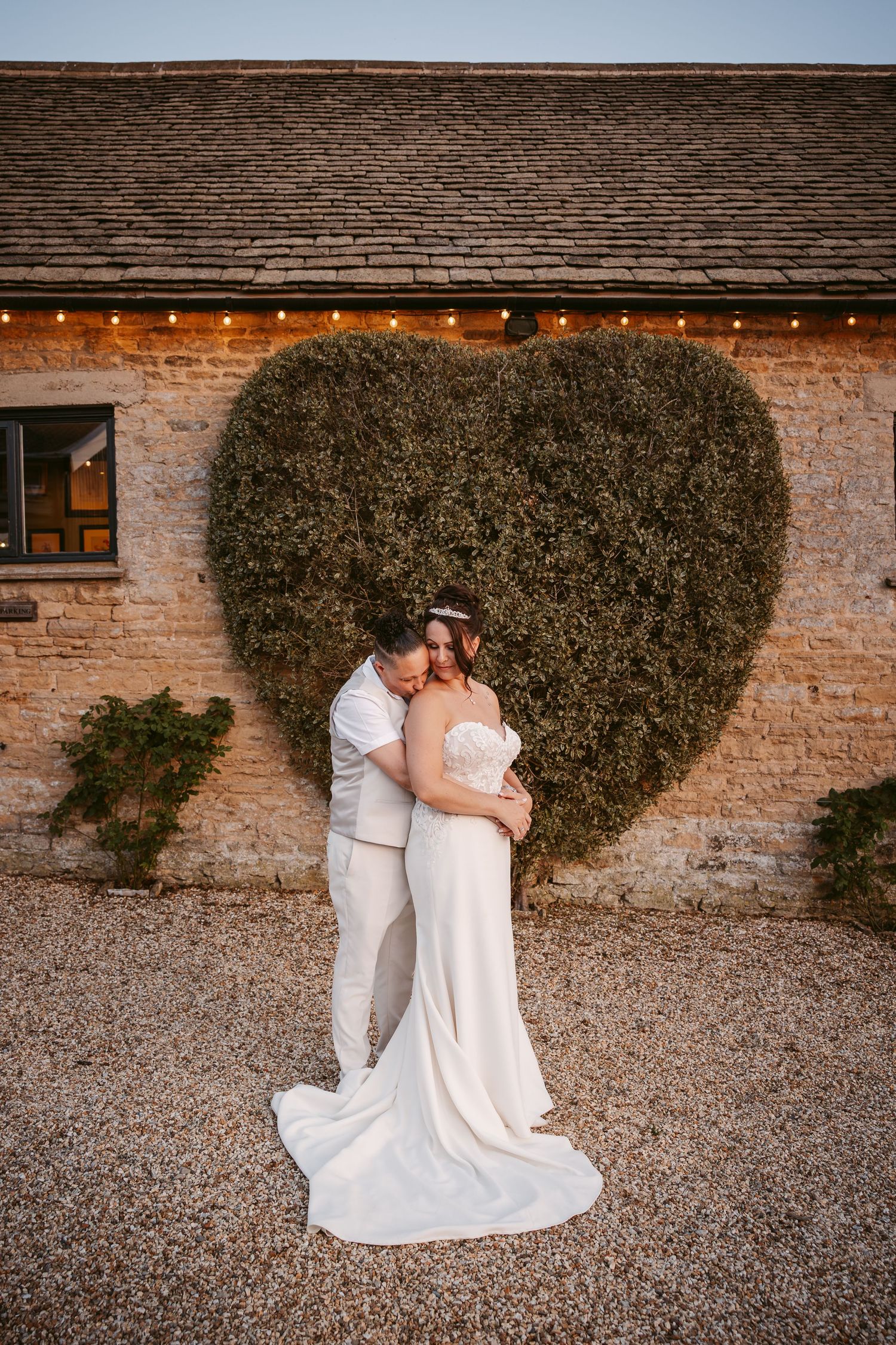 Couple shares intimate moments against rustic brick wall with heart-shaped greenery backdrop.