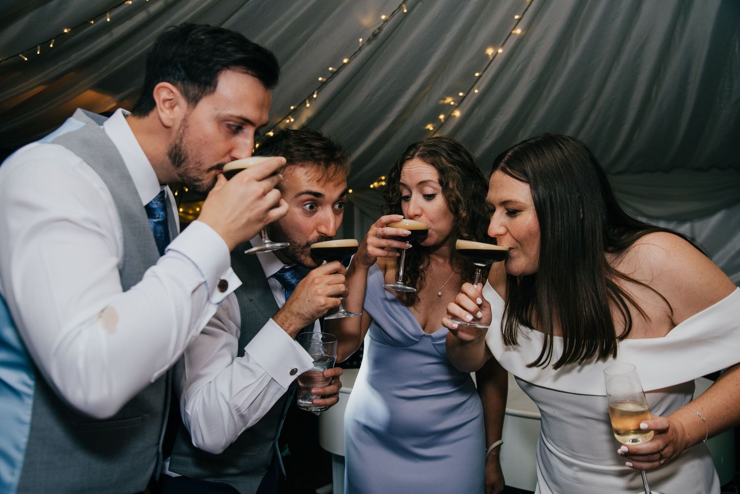 Four wedding guests joyfully sip espresso martinis together at a festive reception under warm fairy lights.