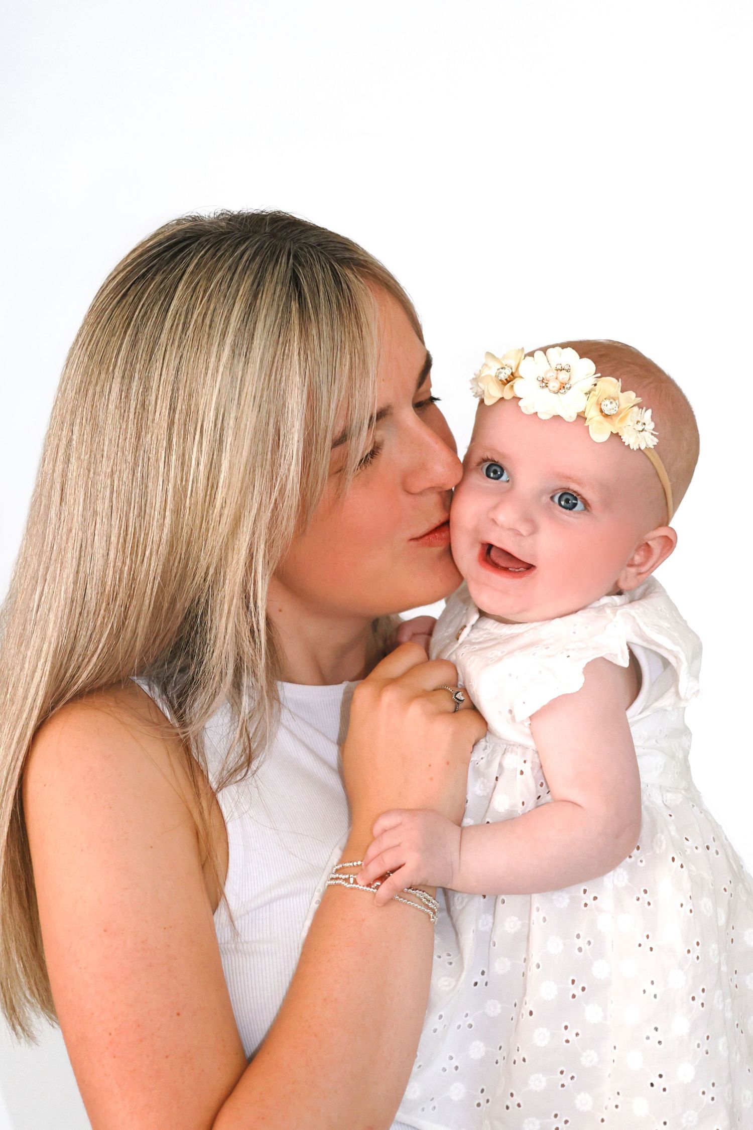 A blonde woman in white dress kissing a smiling baby girl wearing a floral headband and white dress.