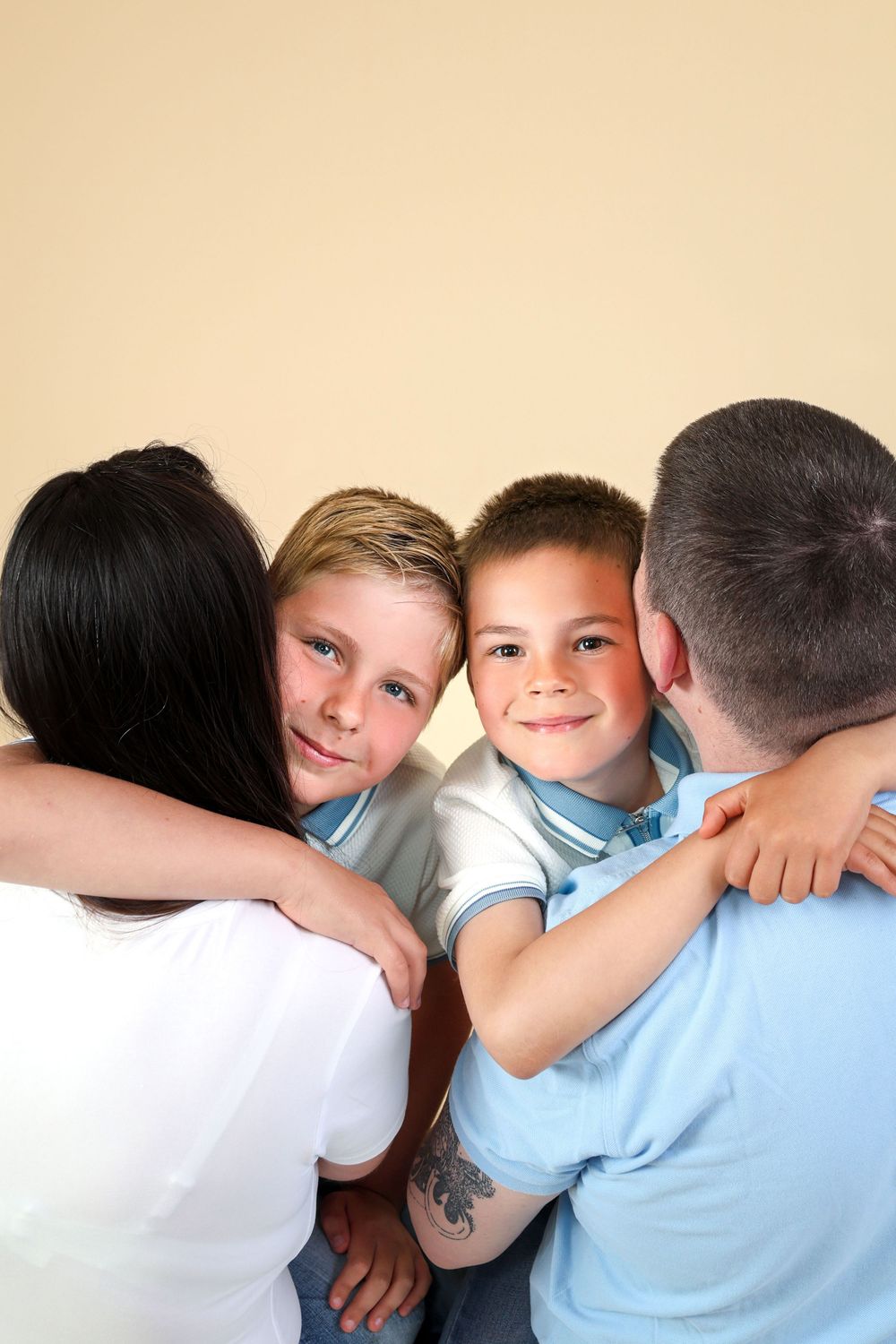 Two happy boys smile while being hugged by their parents in a warm family portrait against a beige background. R.A.M Photography, Stranraer