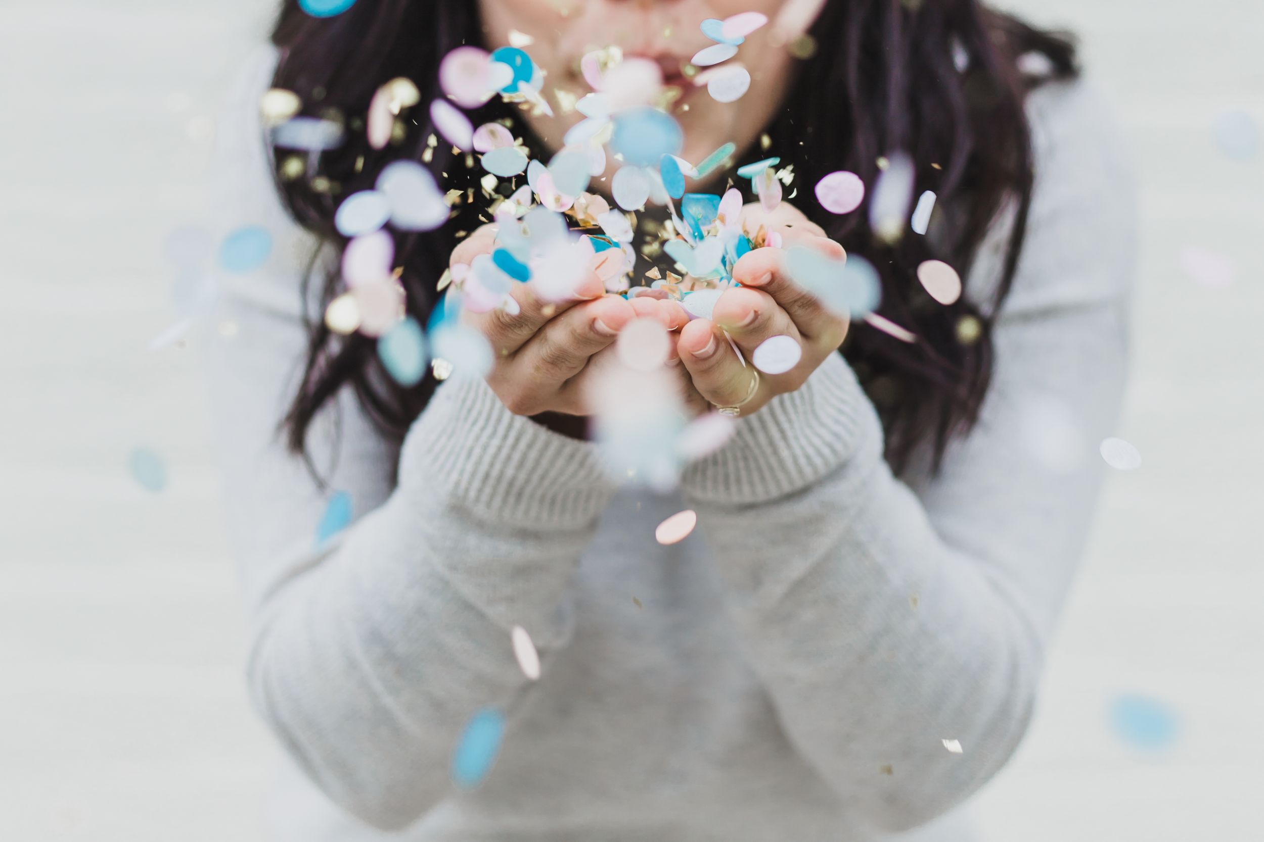 A person in a gray sweater holds their hands up as blue and white confetti falls around them.