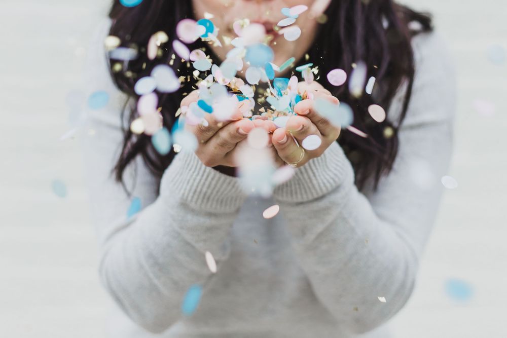 A person in a gray sweater holds their hands up as blue and white confetti falls around them.