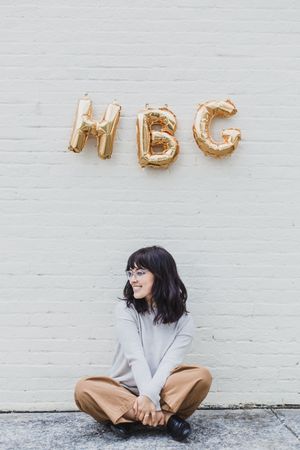 Gold metallic letter balloons spelling out a message against a white brick wall with someone sitting on the ground.