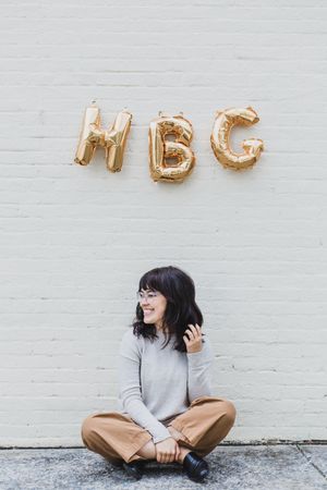 Gold metallic letter balloons displayed on white brick wall with someone sitting and laughing below.
