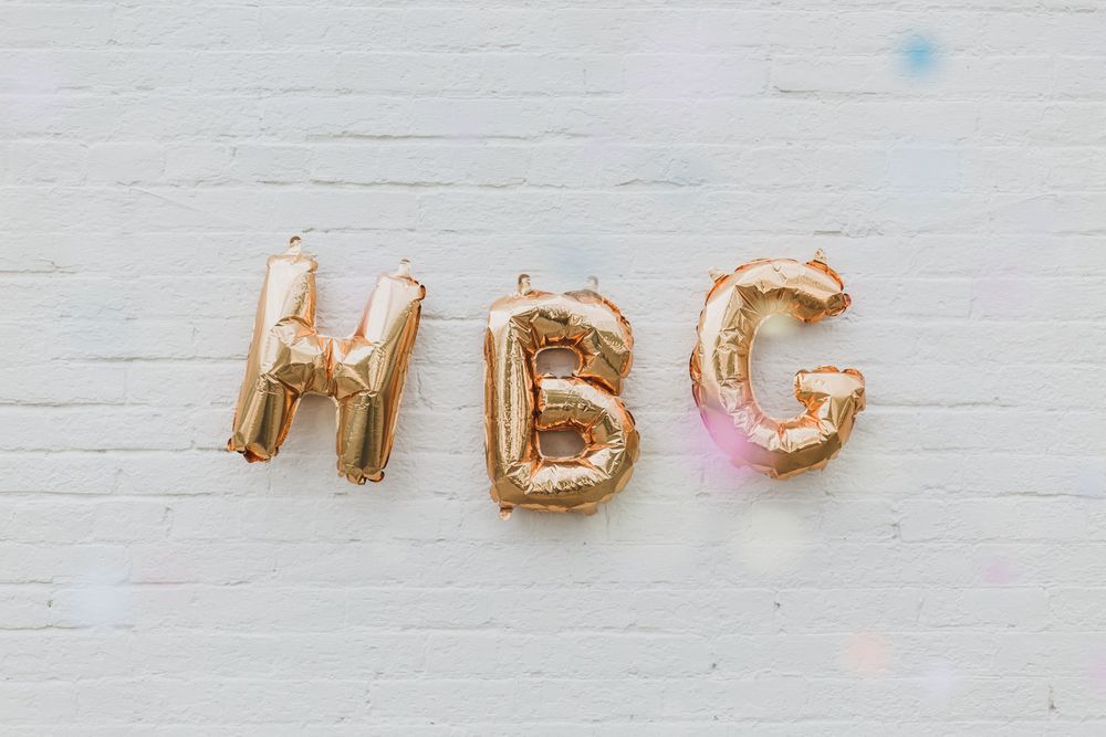 Gold metallic balloon letters spelling out HBG against a white textured wall with soft pastel confetti.
