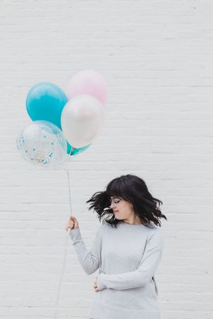 A minimalist photo series shows a person in a light gray sweater holding blue and pink balloons against a white brick wall.