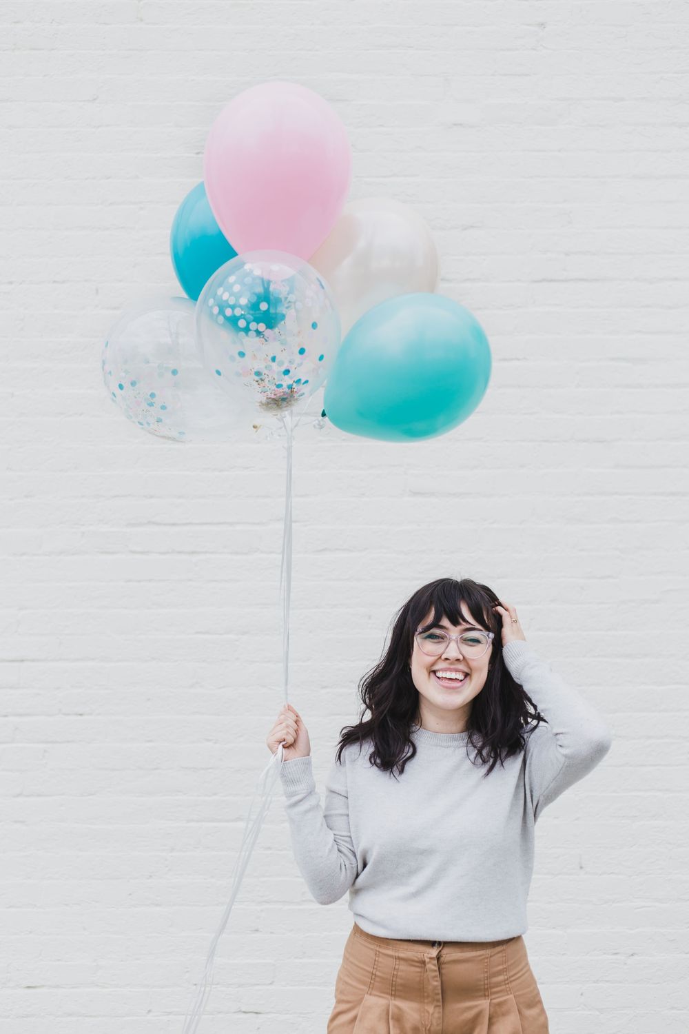 Joyful figure holding pink and turquoise balloons with confetti against white brick wall.