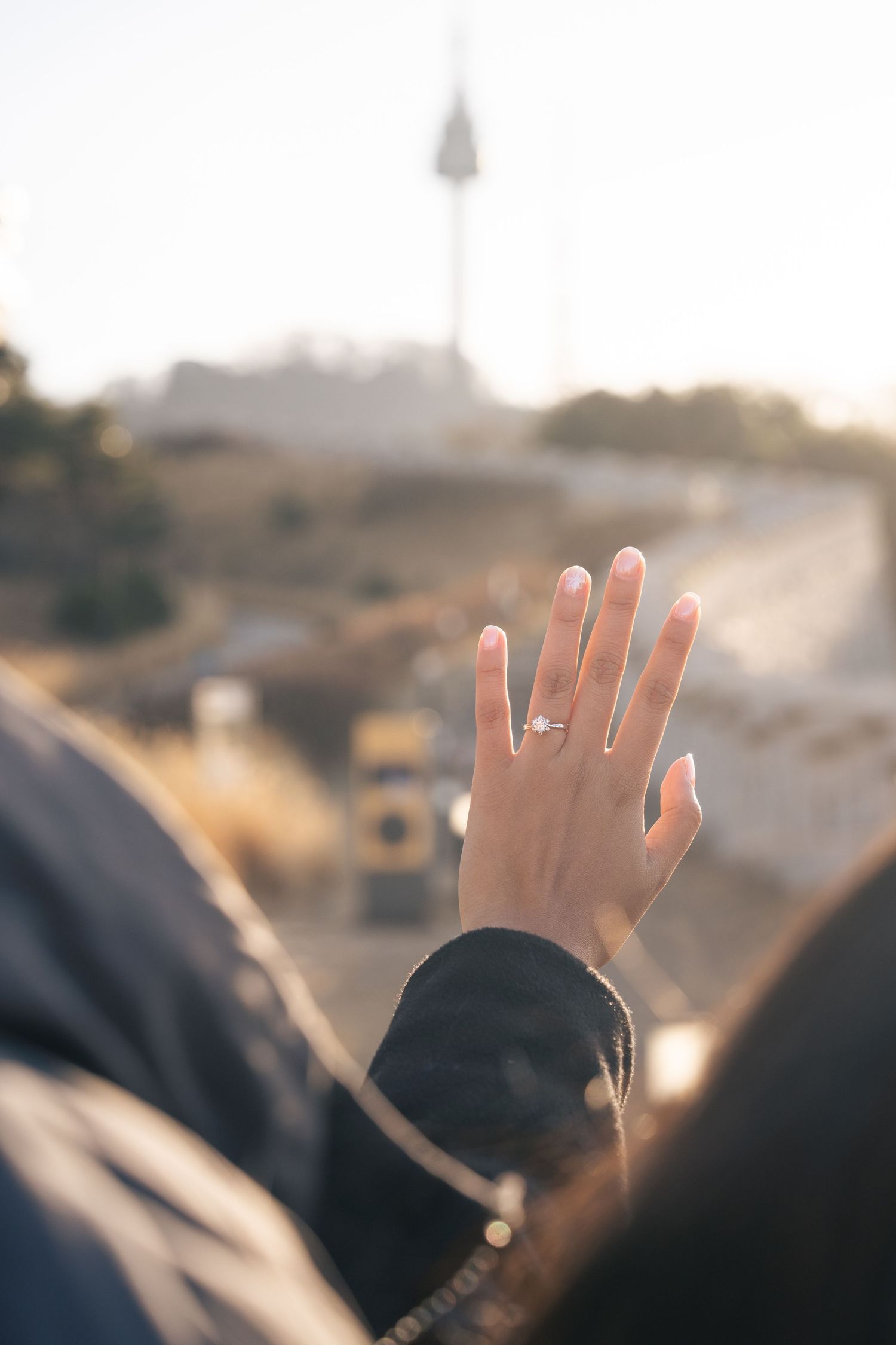 Hand wearing engagement ring raised against blurry sunset background on a road.