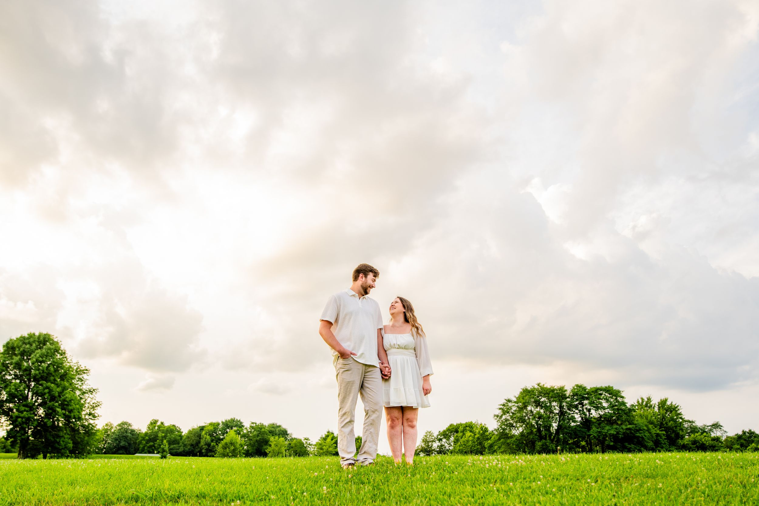 Couple holding hands in a lush green field under dramatic cloudy sky, sharing a romantic outdoor moment.