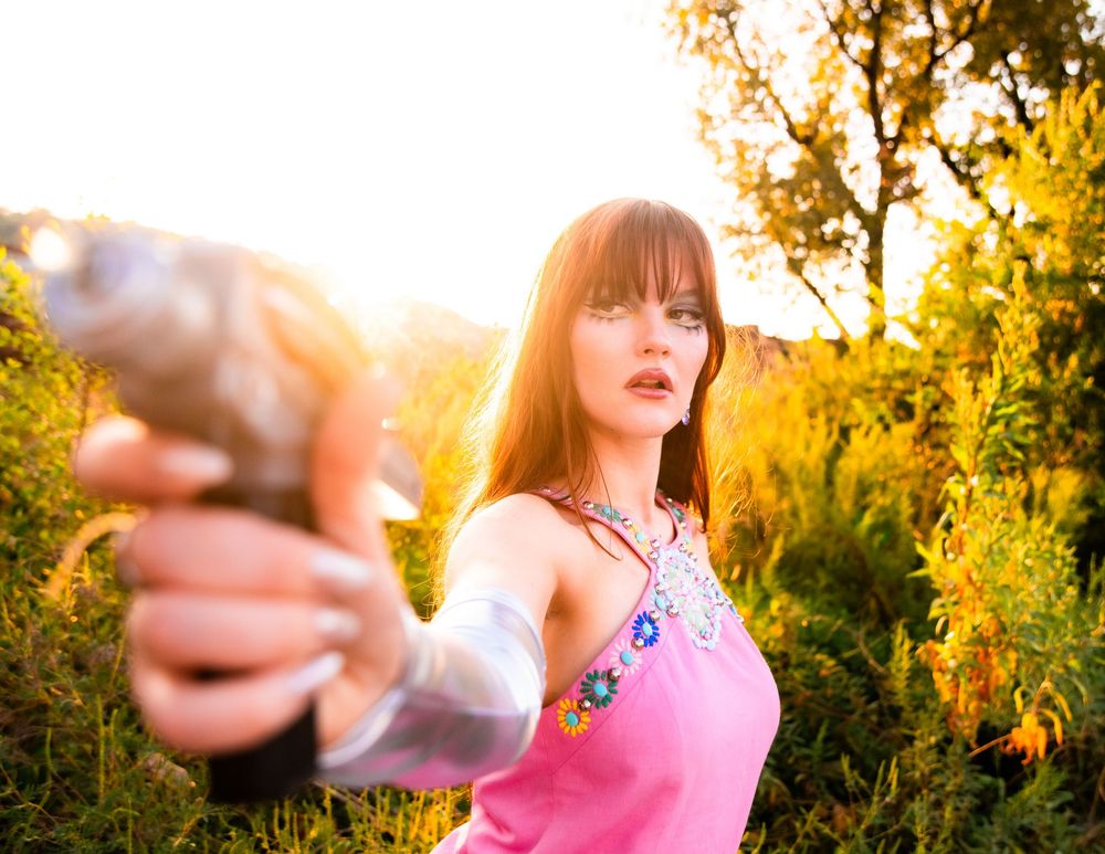 Young woman in pink top pointing gun toward camera in golden sunlight outdoors.
