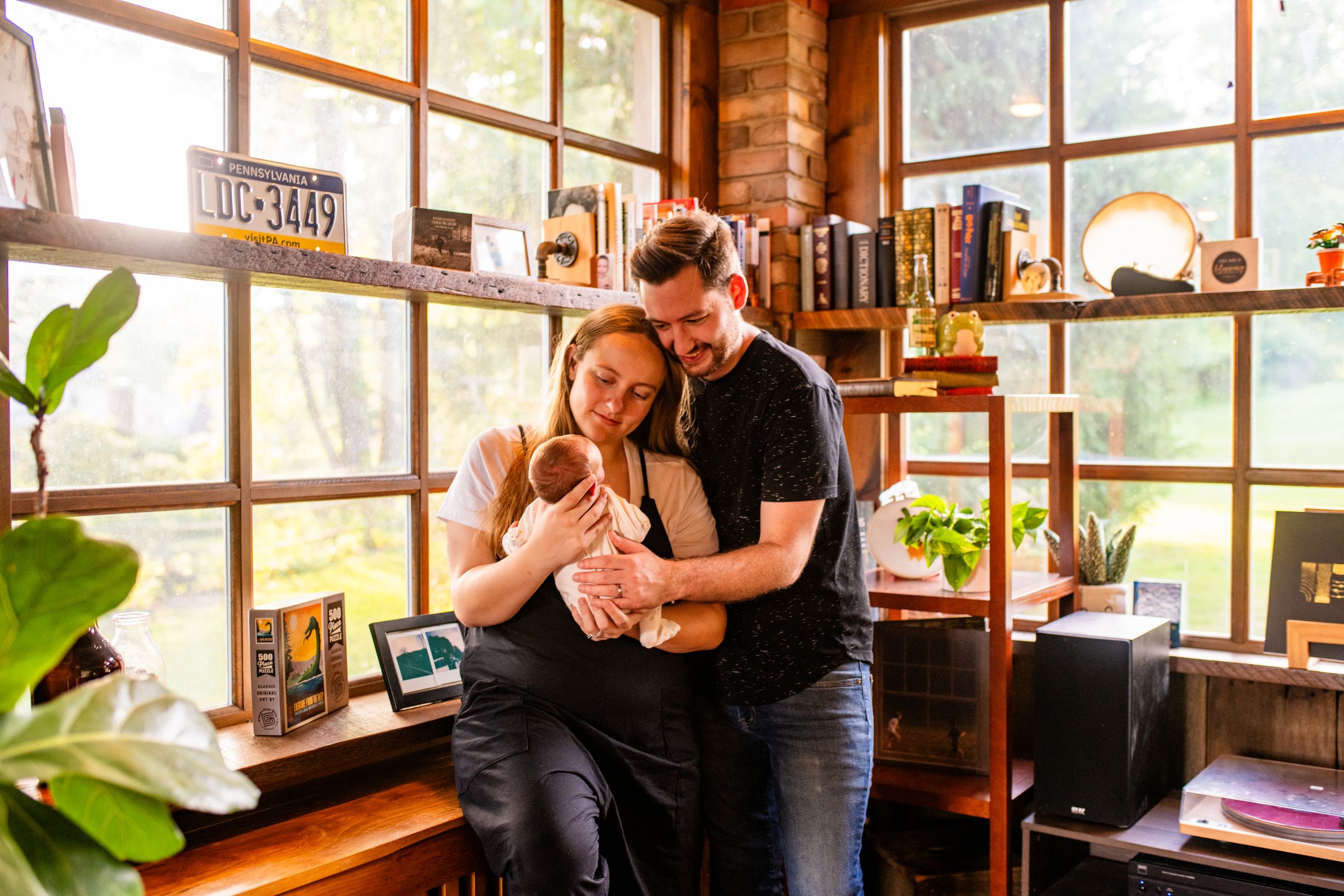 Couple tenderly holds newborn baby in a cozy, book-filled home office with warm natural light.