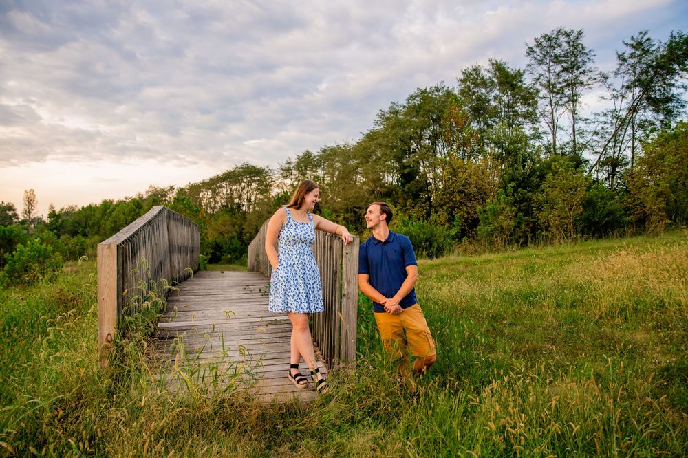 Couple on wooden bridge in green field, man kneeling, woman in blue dress, lush trees and dramatic sky behind them.