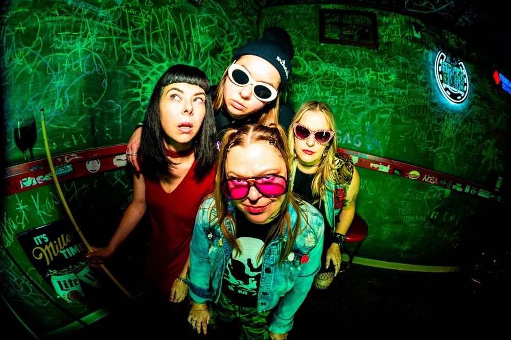 Four young women wearing sunglasses pose together in a vibrant green-lit room at a party venue.