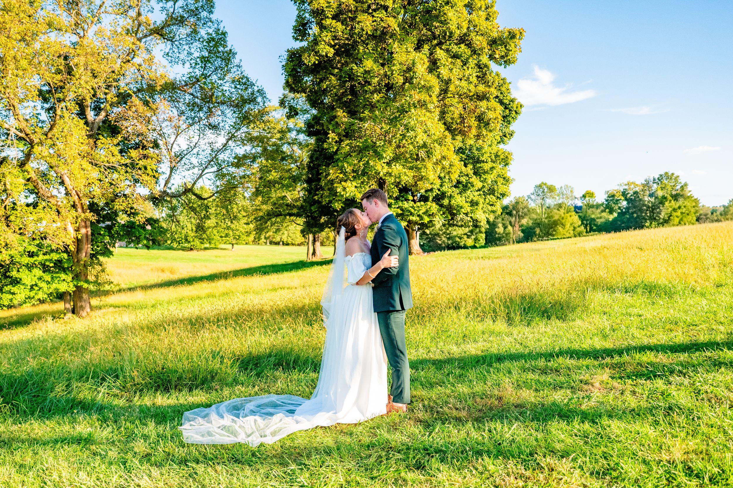 A couple poses in a scenic grassy field with golden sunlight filtering through trees in the background.