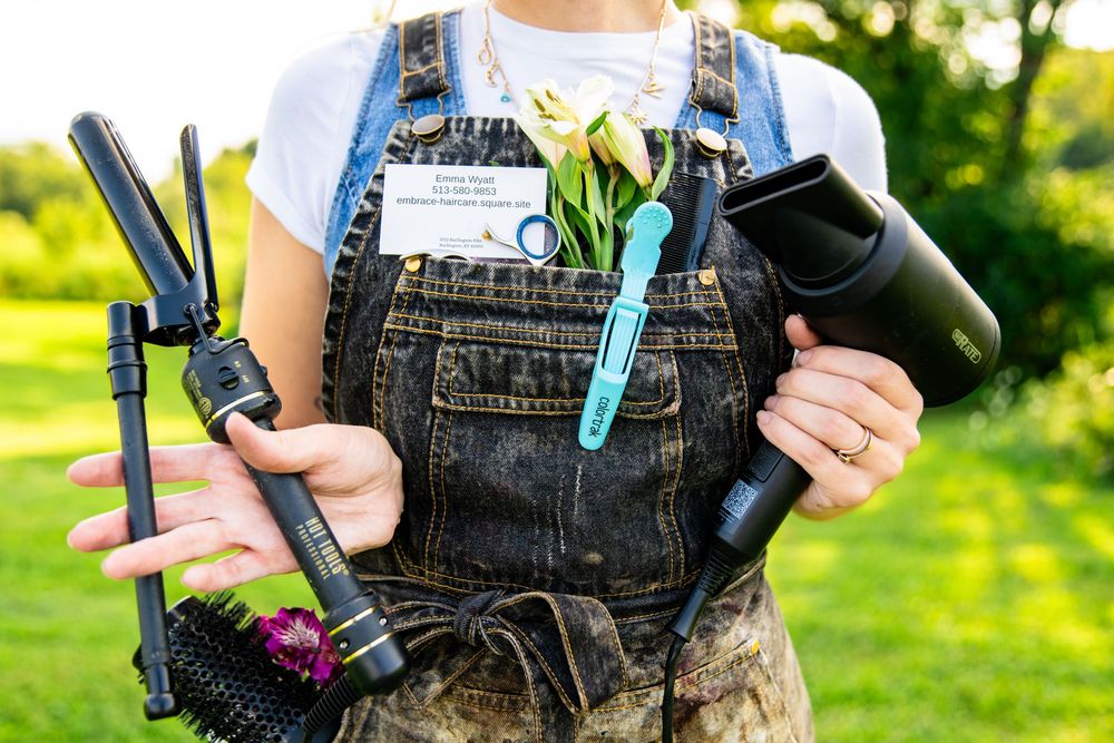 Person in denim overalls holding hair styling tools, scissors, and flowers outdoors.