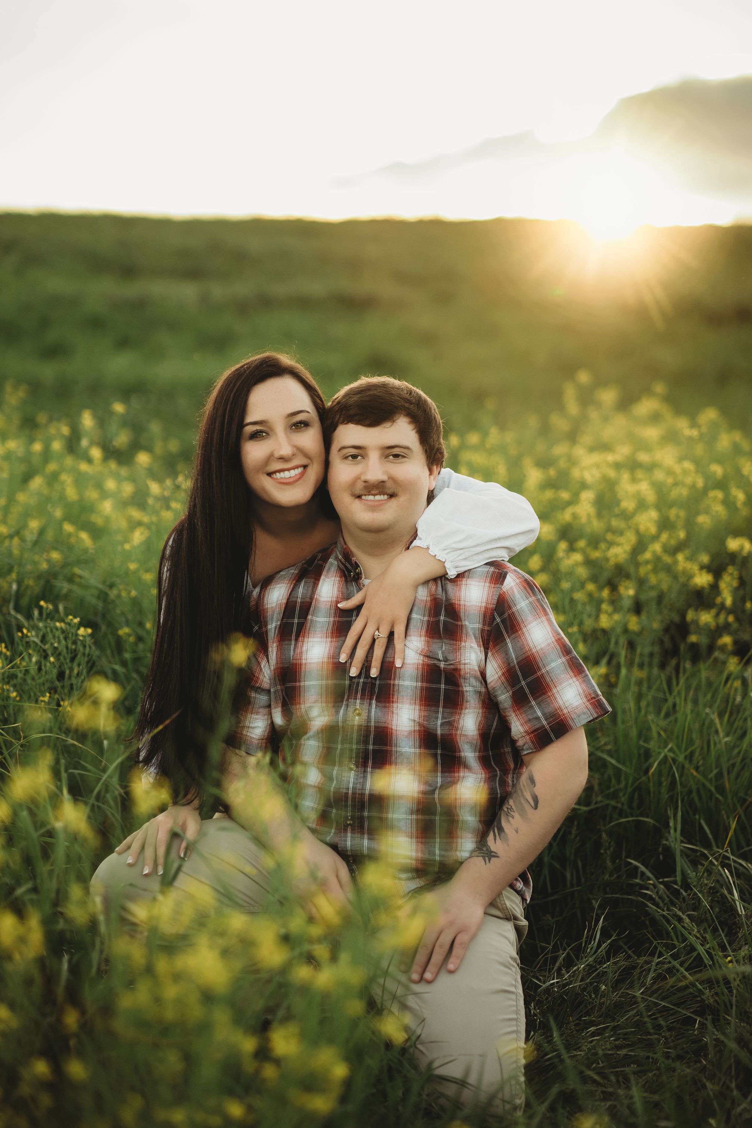Couple smiling in a sunny green field with yellow wildflowers at golden hour sunset.