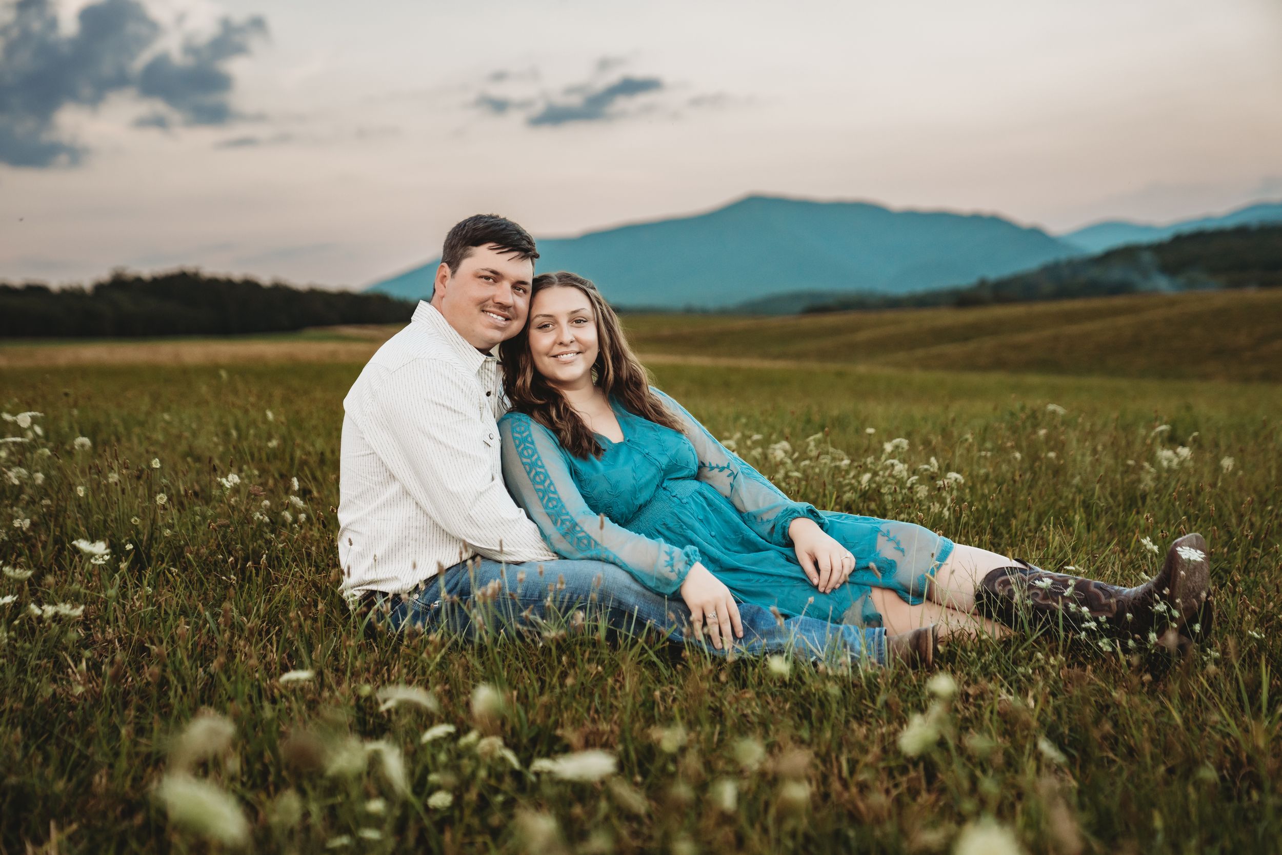 Couple sitting together in a wildflower meadow with mountain views at sunset, woman in teal dress smiling.