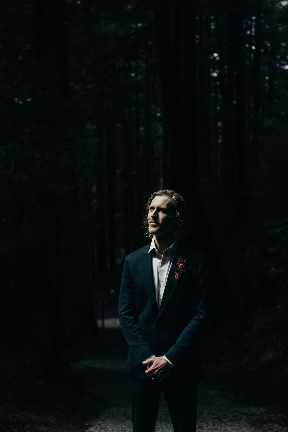 Dark moody forest wedding portrait with groom in black suit against evergreen trees.