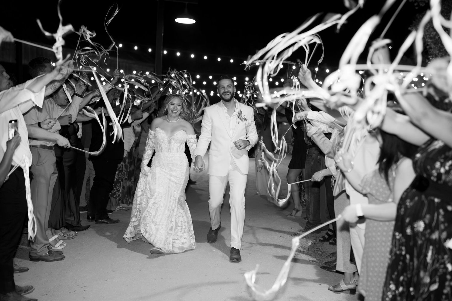 A large group of wedding guests dance together under strings of lights at an outdoor evening reception celebration.