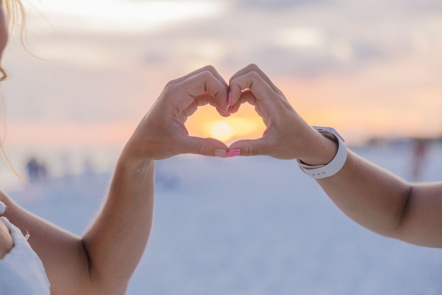 Hands form a heart shape silhouetted against a vibrant sunset sky over water.