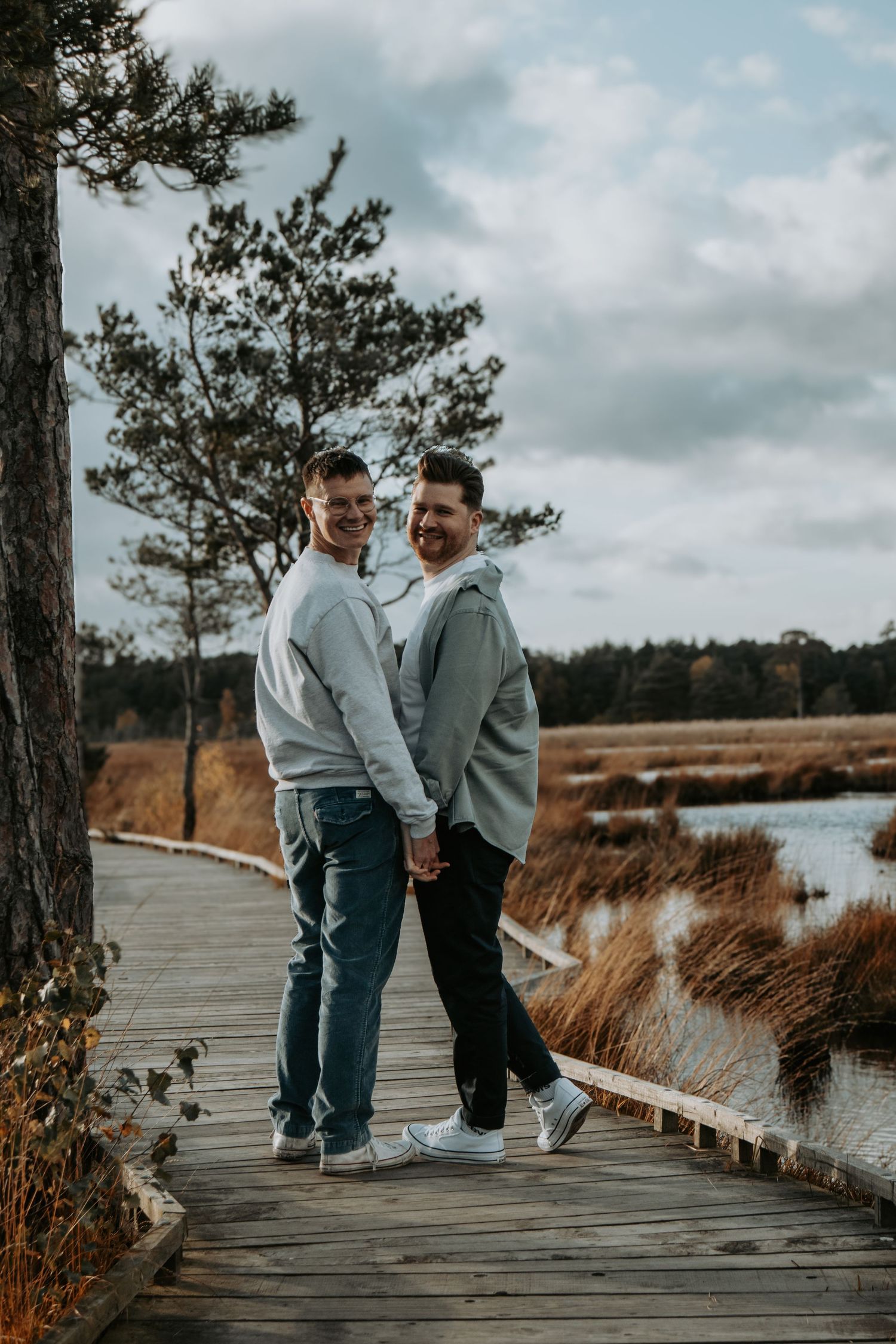 Two people in matching gray sweaters walk together on a wooden boardwalk by a scenic lake on a cloudy day.