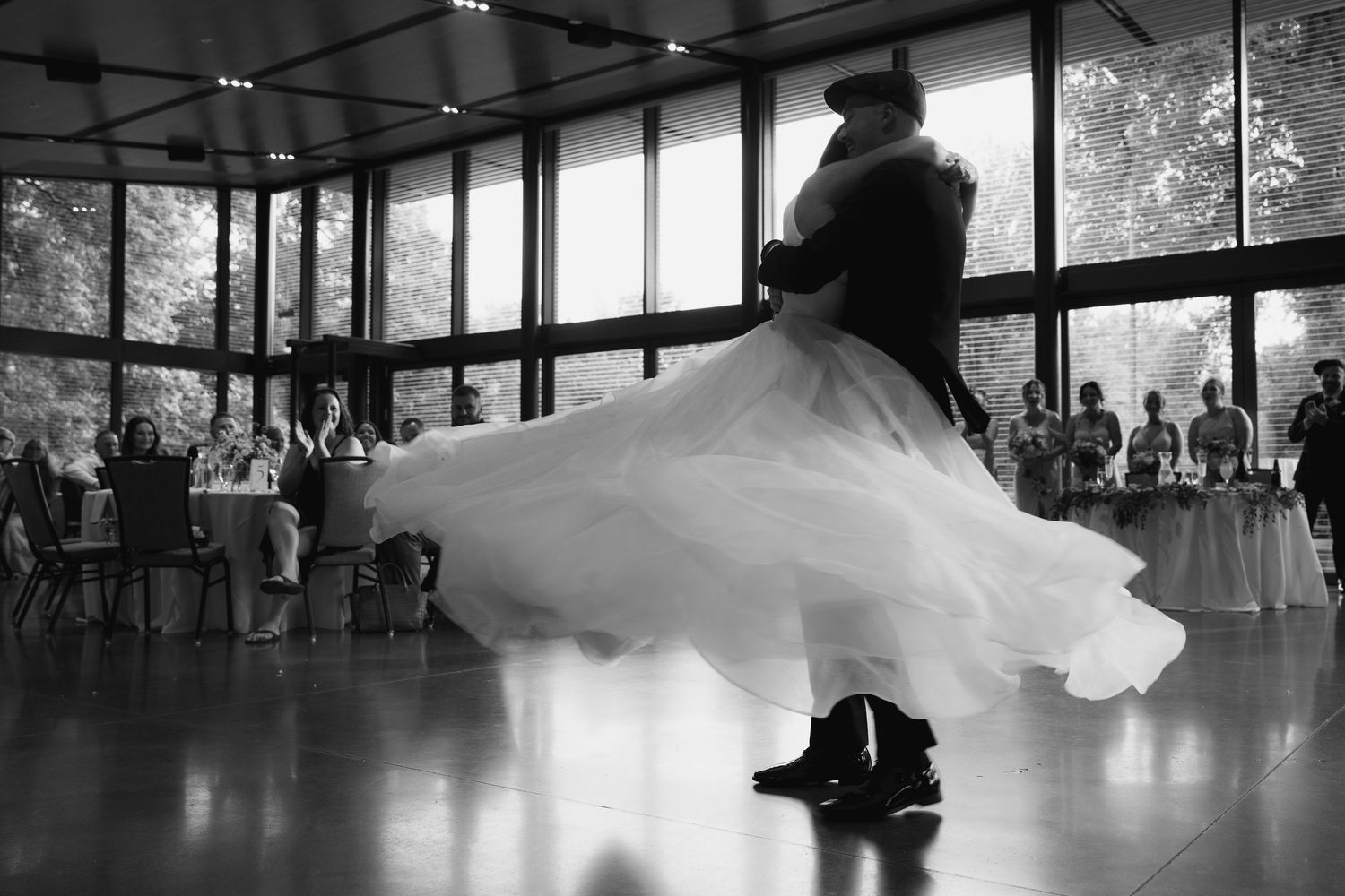 Dancers in formal attire perform elegant movements in a bright dance studio with floor-to-ceiling windows.