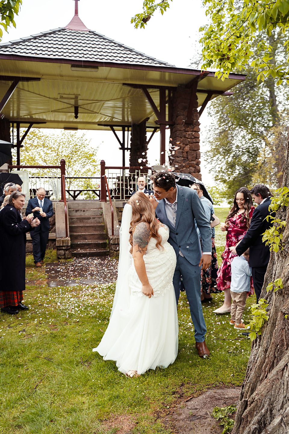 A couple shares their first kiss as newlyweds under a gazebo while guests shower them with flower petals in celebration.