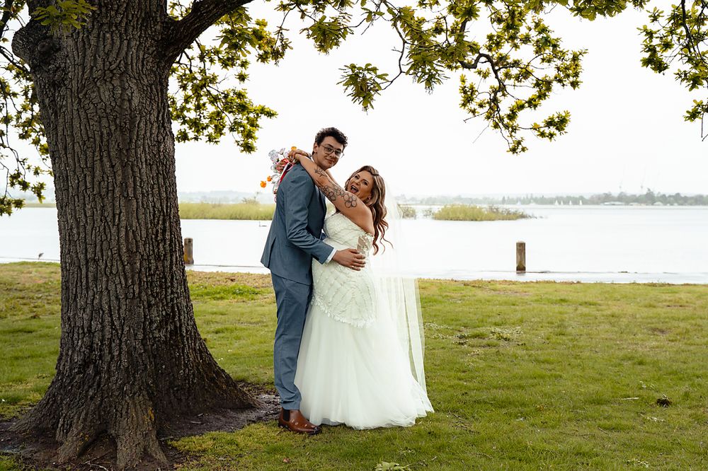 A couple in wedding attire poses together by a large tree near a serene lakefront on a sunny day.