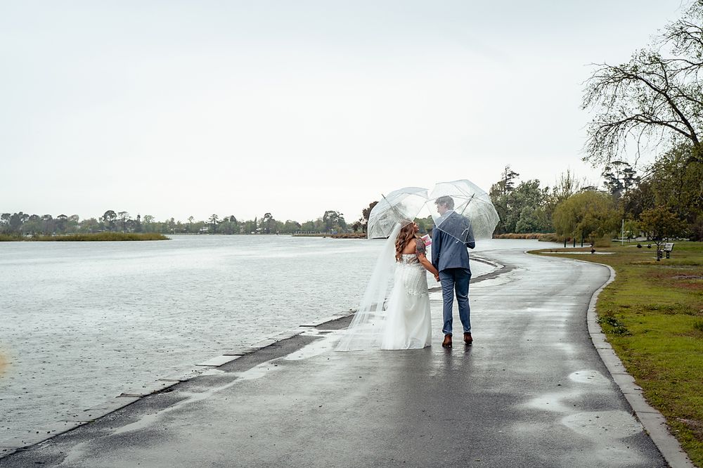 A couple shares an umbrella while walking along a rainy lakeside path during their wedding day.