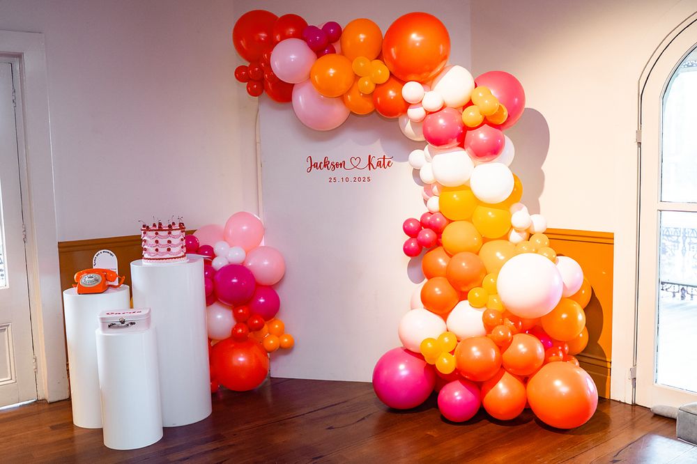 A balloon arch installation in pink, orange, and white colors decorates a corner with white pedestals against a white wall.