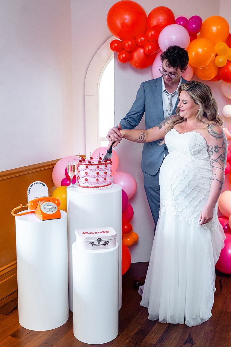 A couple cuts their wedding cake together surrounded by colorful orange and pink balloon decorations at their celebration.