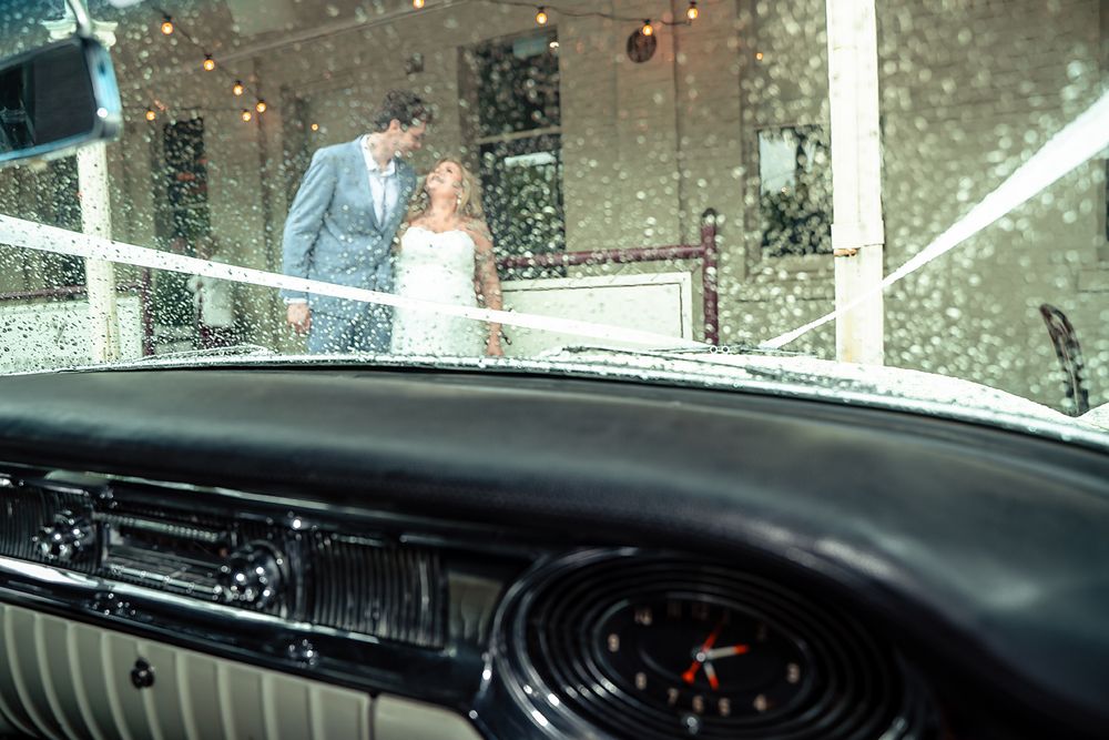 A romantic wedding scene viewed through a vintage car windshield with raindrops on a rainy day.