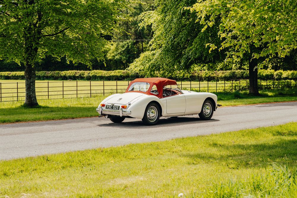 Vintage white sports car with red top driving on a scenic tree-lined country road.