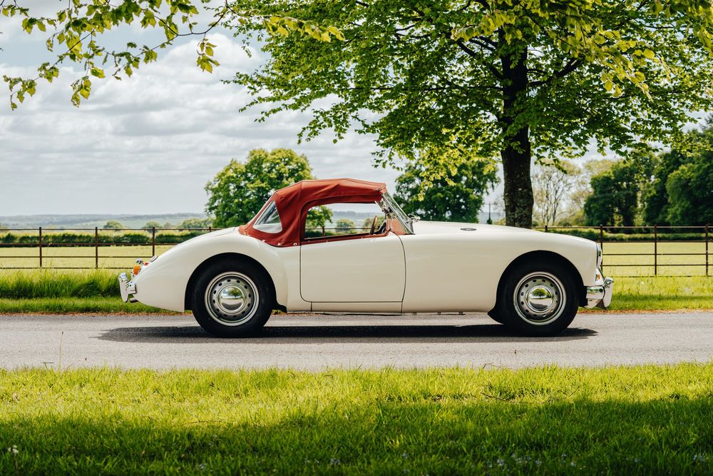 White vintage sports car with red roof shown in profile view against lush green landscape.
