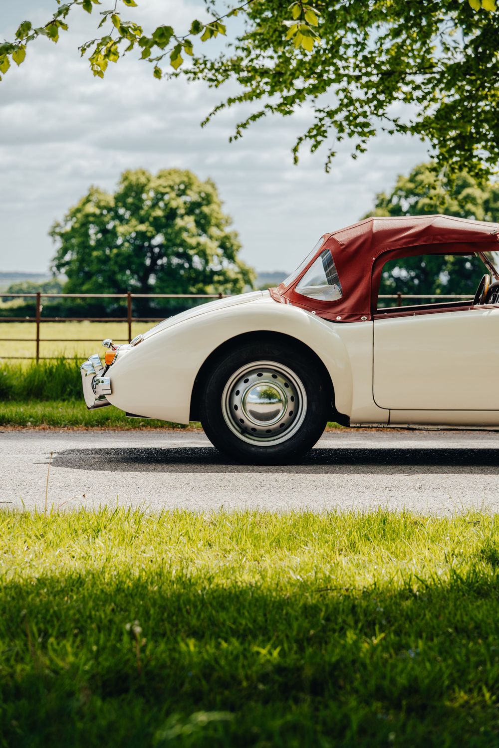 Side view of a classic white and red vintage car parked on a country road with green trees in the background.