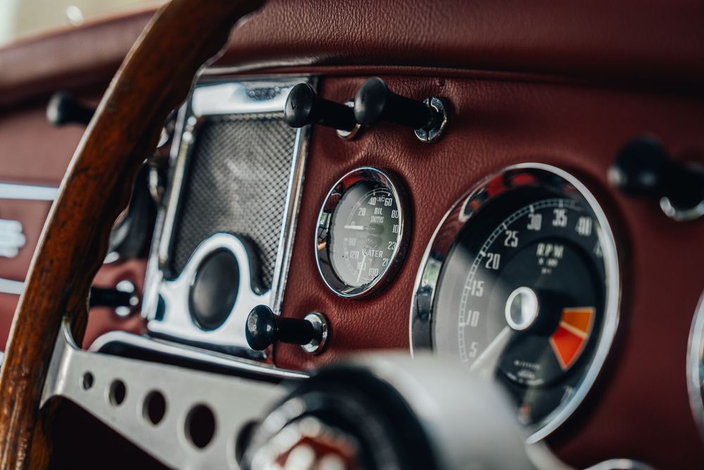 Classic car dashboard showing vintage gauges, controls, and burgundy leather interior details in warm lighting.