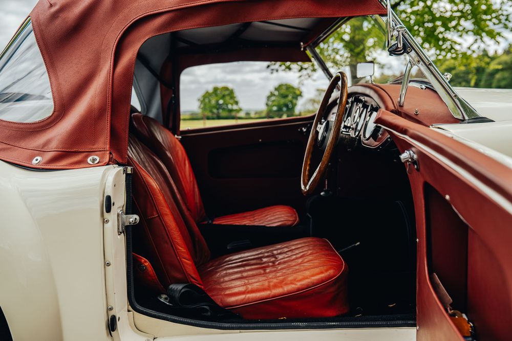 Interior view of vintage car showing red leather seats and wooden steering wheel with convertible top visible.