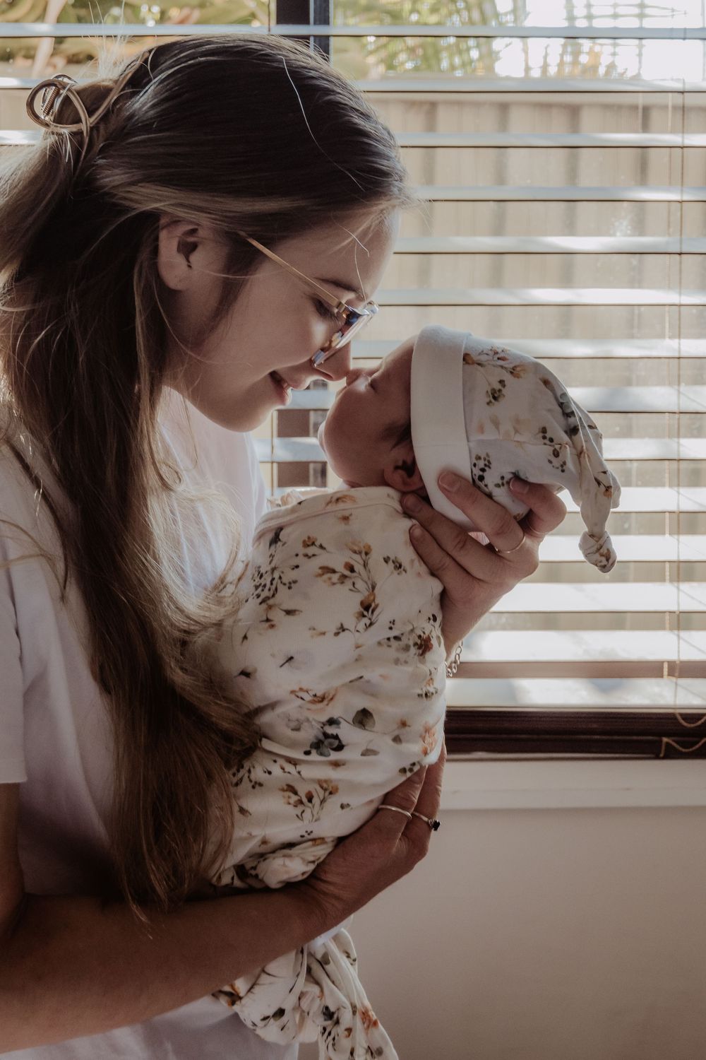 Intimate portrait by window of caregiver holding baby in floral outfit at a central coast newborn photoshoot.