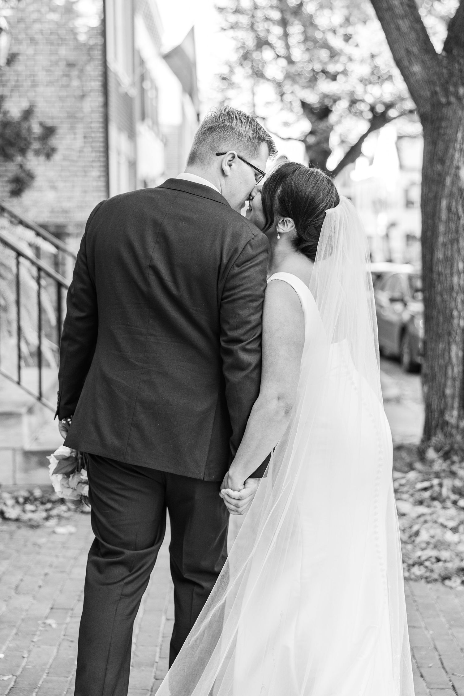 Black and white wedding photo showing couple in formal attire standing together on brick sidewalk with flowing veil.
