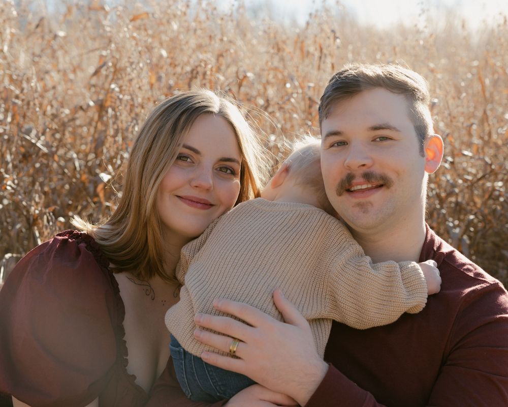 A couple in burgundy sweaters smiles while holding their baby outdoors with golden autumn grass in the background.