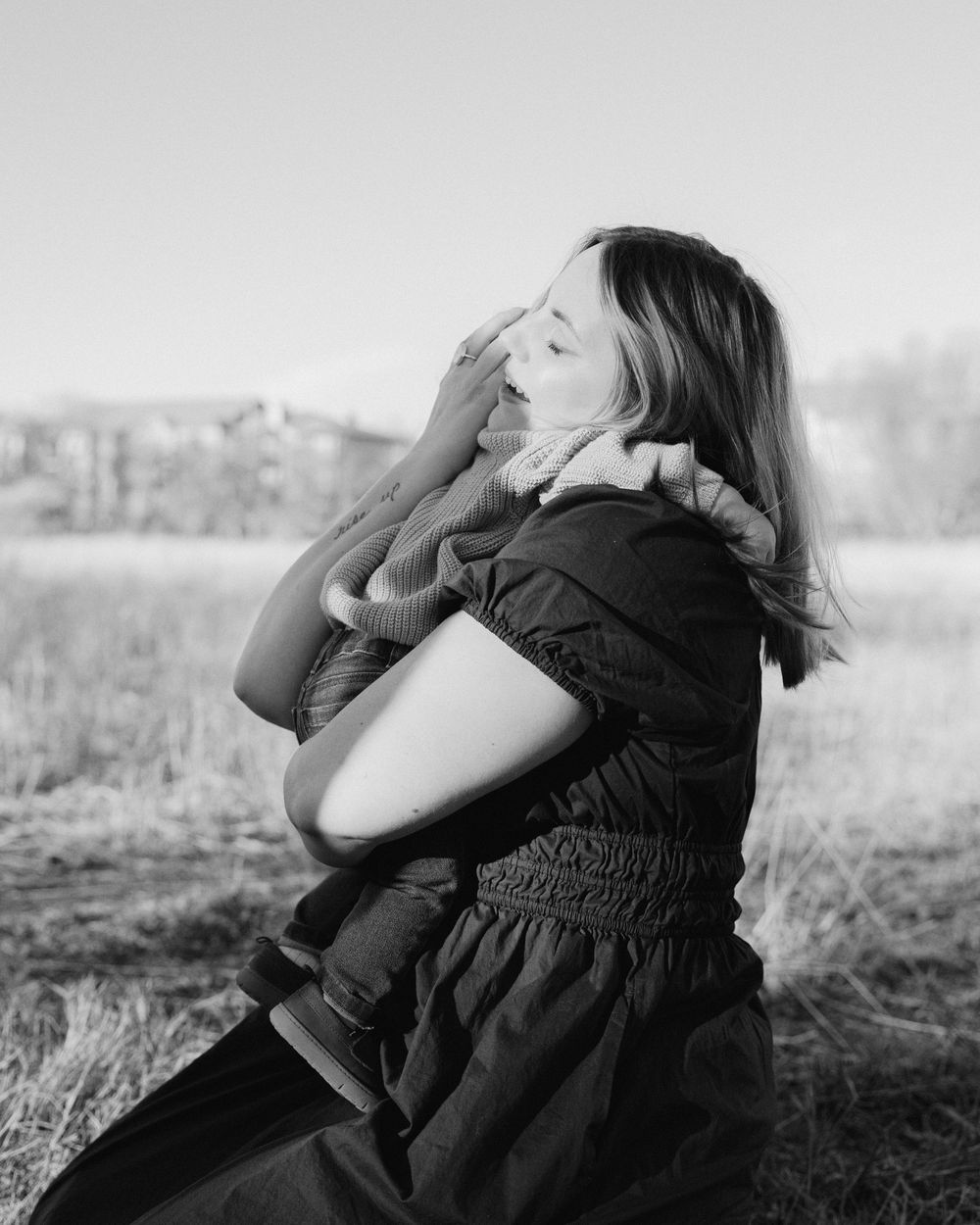 Black and white portrait of someone in a dark dress sitting outdoors with eyes closed and hand raised in sunlight.