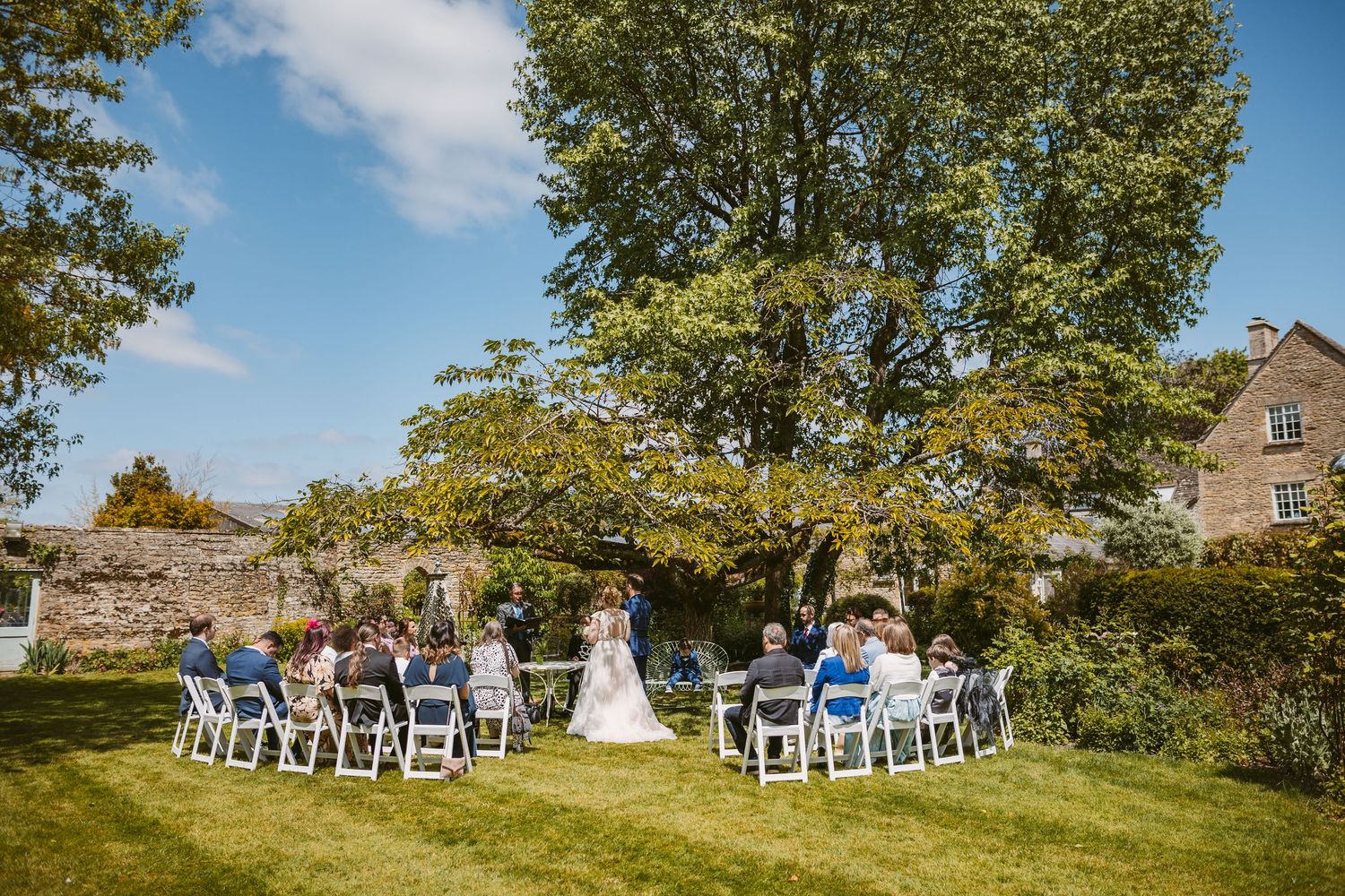 Panoramic view of an outdoor wedding ceremony setup with white chairs on a grassy hill under tall trees on a sunny day.