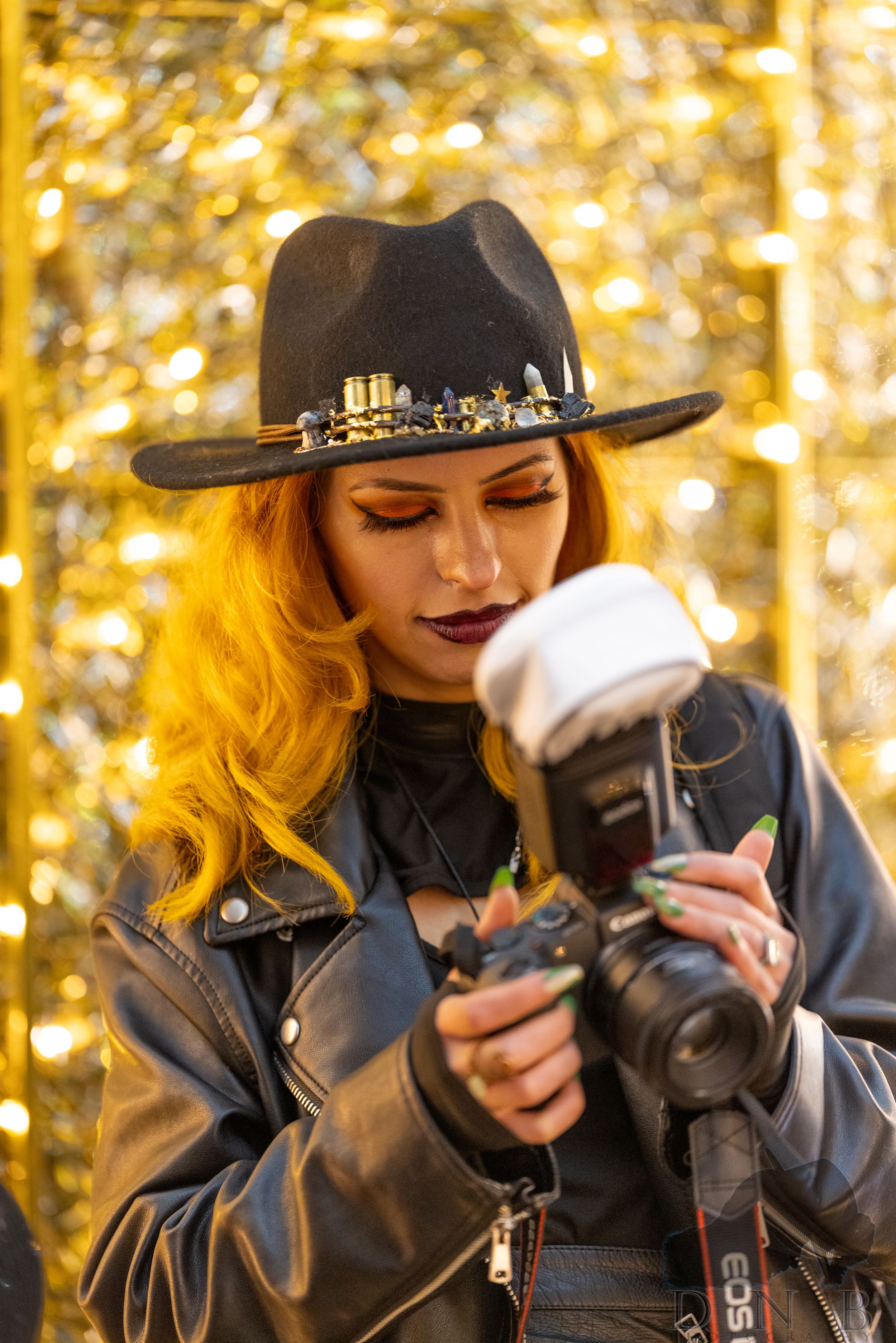 A woman with orange hair and a decorated hat adjusts her Canon camera with flash.